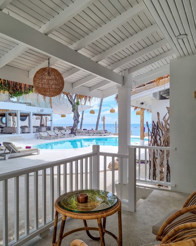 A serene tropical resort scene featuring a covered patio with wicker furniture overlooking a sparkling pool and beach, ideal for relaxing vacations in Thailand.