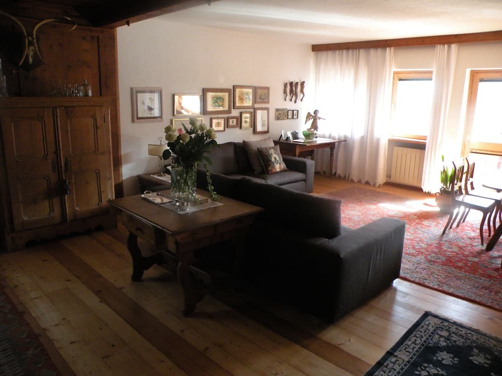 A cozy living room with wooden floors, rustic furniture, and warm lighting creates an inviting atmosphere perfect for a relaxing getaway, featuring a central coffee table adorned with flowers and framed artwork on the walls.