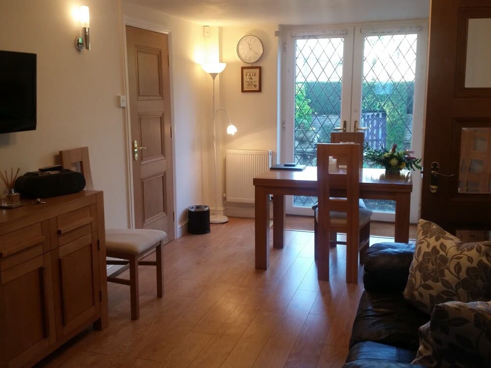 A cozy living room with wooden flooring, modern furniture, and a view of greenery through glass doors, ideal for travelers seeking a comfortable and inviting accommodation.
