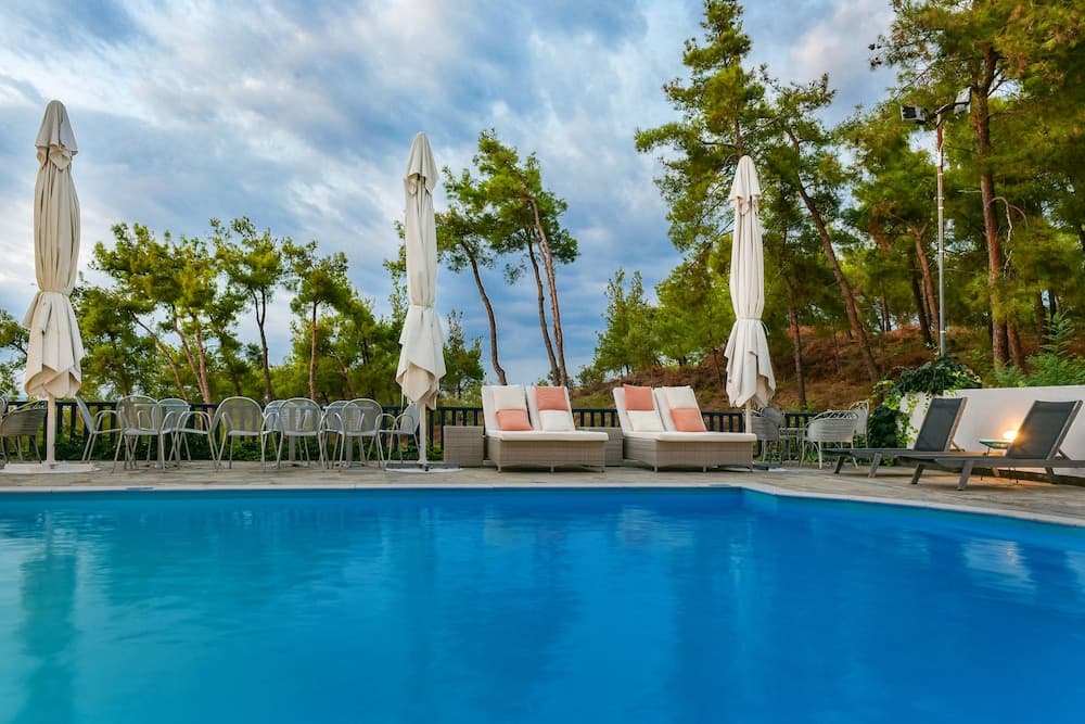 A serene resort poolside scene features vibrant blue water, cozy lounge chairs with pink cushions, closed white umbrellas, and lush green trees under a partly cloudy sky, ideal for relaxation and leisure travel.