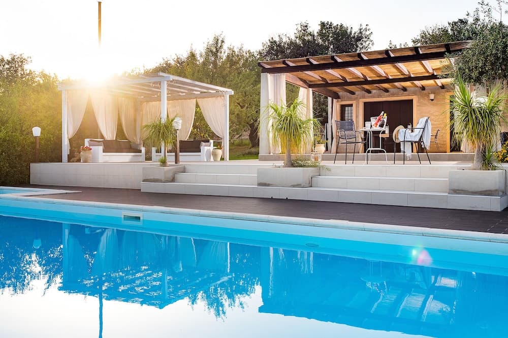 A serene poolside retreat features a tranquil blue pool, a cozy pergola with curtains, and a shaded dining area, perfect for a relaxing vacation in a lush, sunlit garden setting.