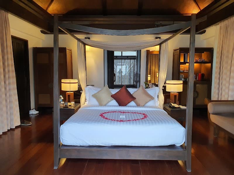 A cozy bedroom at the Nora Buri Resort & Spa on Koh Samui features a four-poster bed adorned with a heart-shaped petal arrangement, warm wooden floors, and ambient lighting, creating an inviting and romantic atmosphere ideal for travelers seeking relaxation and luxury.