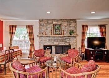 A cozy living room with a stone fireplace, patterned armchairs, and large windows draped in red curtains creates an inviting atmosphere ideal for family gatherings or a relaxing retreat.