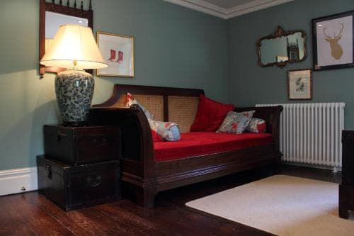 A cozy bedroom with teal walls, a red cushioned daybed adorned with pillows, vintage decor including framed artwork and mirrors, and warm lighting creates an inviting atmosphere ideal for a relaxing getaway.