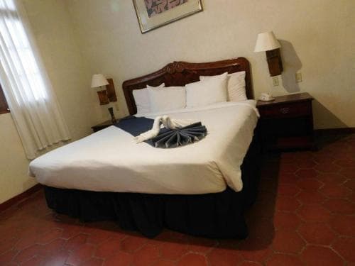 A cozy hotel room features a neatly made bed adorned with a decorative towel swan, warm terracotta flooring, and classic wooden furnishings, ideal for travelers seeking comfort and charm.
