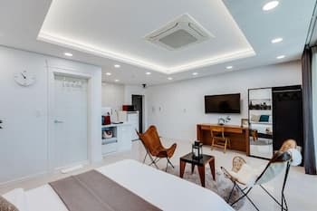 A cozy modern studio apartment features a minimalist design with a white bed, leather chair, wooden desk, and flat-screen TV, creating an inviting atmosphere ideal for travelers seeking comfort and style in urban accommodations.