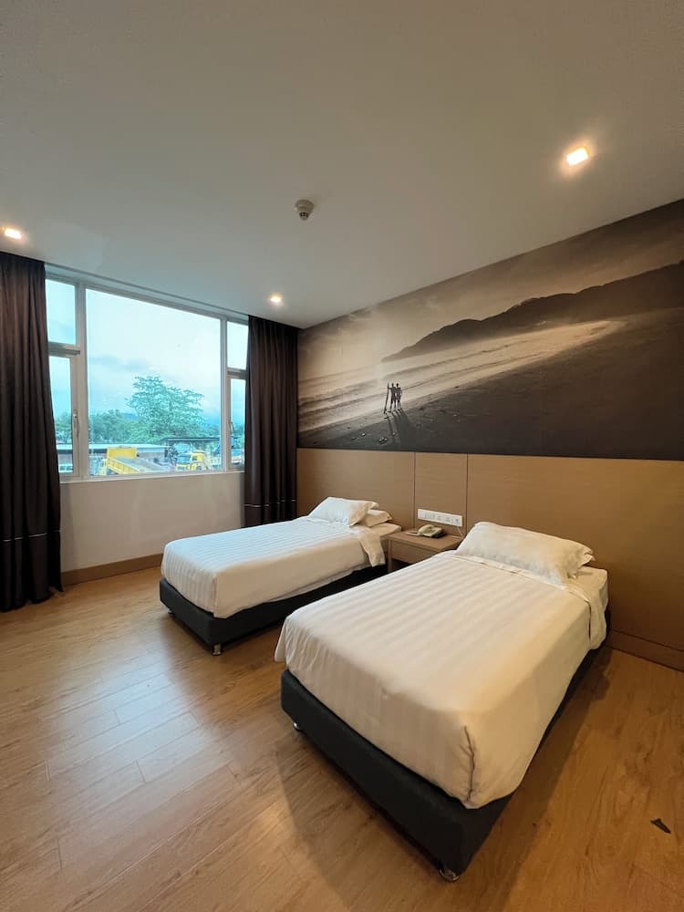 A cozy hotel room with two neatly made beds, a beach mural on the wall, wooden flooring, and a window offering a serene outdoor view, ideal for travelers seeking comfort and relaxation.
