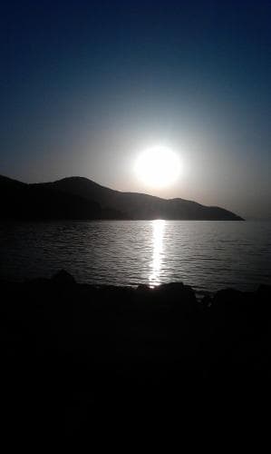 A serene sunset over calm waters reflects off the surface near silhouetted hills, creating a tranquil coastal scene ideal for relaxation and photography.