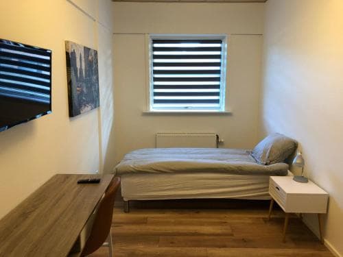 A cozy minimalist bedroom with a neatly made bed, wooden desk, flat-screen TV, and window with blinds, ideal for travelers seeking a simple and functional accommodation space.