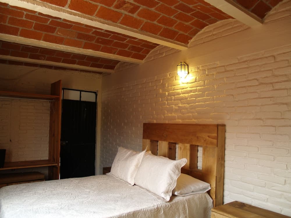 A cozy bedroom with exposed brick walls and ceiling beams exudes rustic charm, featuring a wooden bed, soft lighting from a wall-mounted lamp, and a quaint black door, ideal for travelers seeking a serene retreat.
