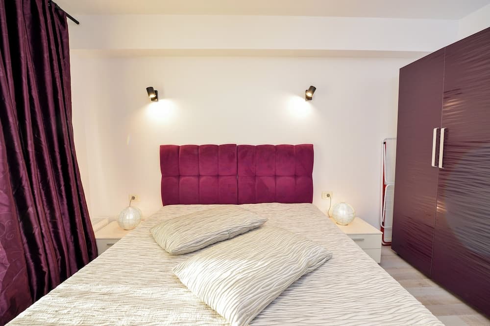 A cozy bedroom features a plush bed with a vibrant purple headboard, matching curtains, and ambient lighting, ideal for travelers seeking comfort and style in their accommodations.