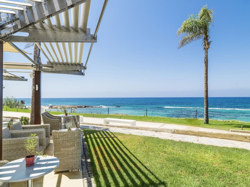 A serene coastal retreat features wicker seating under a modern pergola, casting shadows on lush green grass beside a tranquil ocean view, ideal for relaxing beachside vacations.