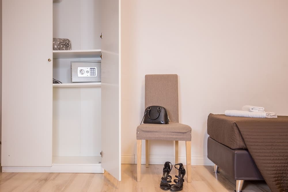 A cozy hotel room corner features a neatly arranged bed, a chair with a handbag, sandals on the floor, and an open wardrobe with a safe, ideal for travelers seeking comfort and security.