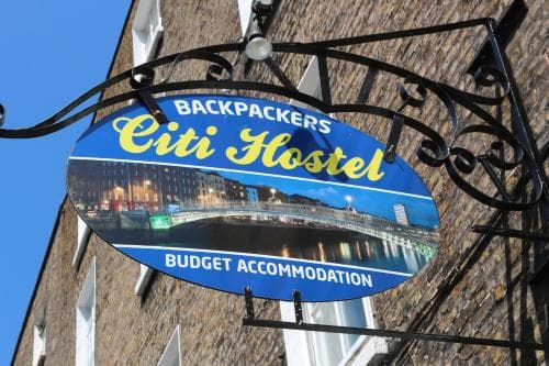 A vibrant blue sign for "Backpackers Citi Hostel" hangs against a brick building under a clear sky, inviting travelers seeking budget accommodation in an urban setting.