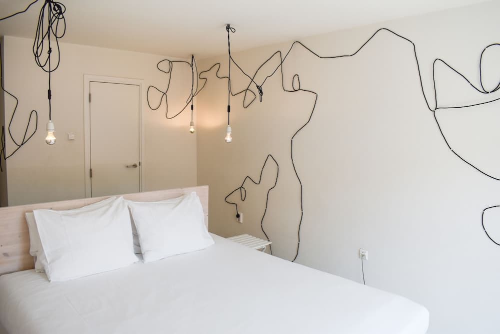 A minimalist bedroom features an artistic wire outline of mountains and animals on the wall, paired with simple hanging light bulbs, creating a serene and modern atmosphere ideal for travelers seeking unique accommodations.