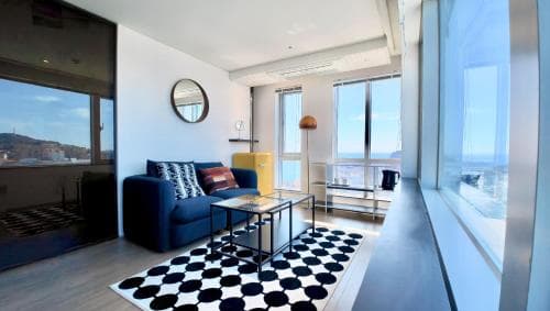 A modern apartment with a cozy living area featuring a blue sofa, checkered rug, and large windows offering scenic views, ideal for travelers seeking comfort and style in an urban retreat.