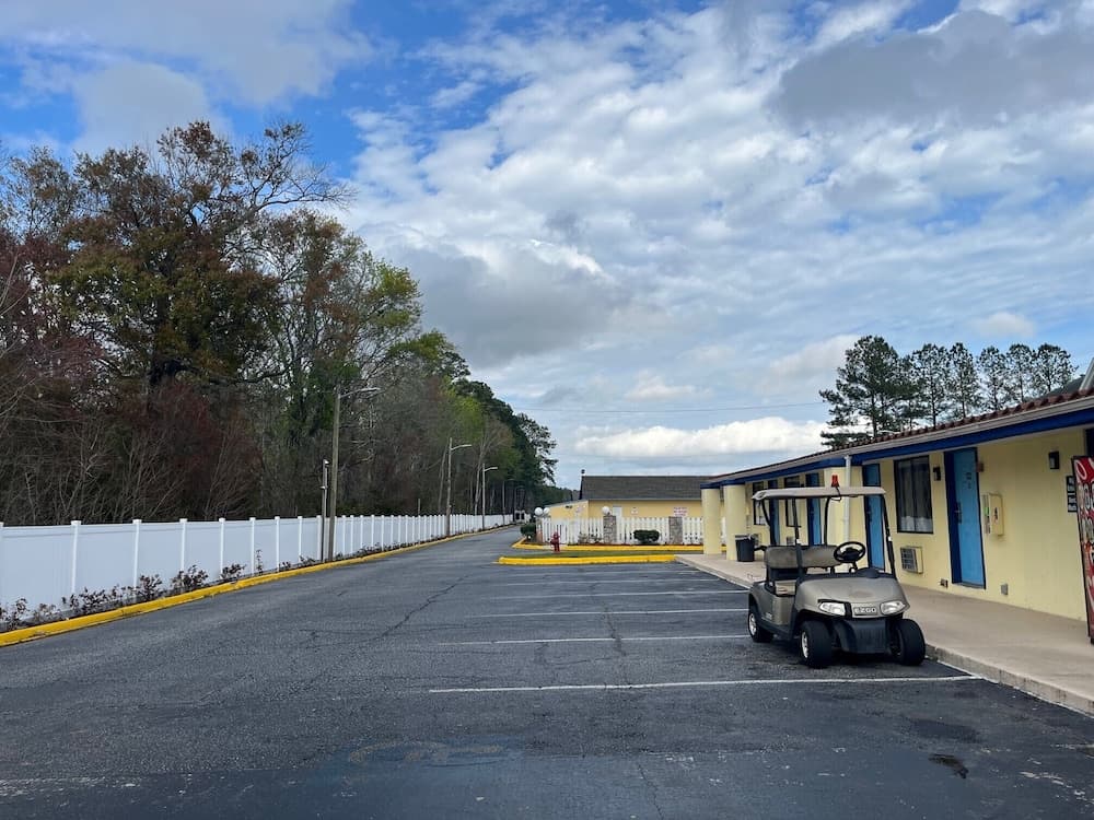A serene motel parking lot features a parked golf cart under a partly cloudy sky, surrounded by lush trees and a white fence, ideal for travelers seeking a quiet retreat.