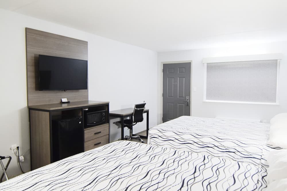 A minimalist hotel room features a neatly made bed with patterned sheets, a mounted TV, a small desk with a chair, and a compact kitchenette, ideal for travelers seeking comfort and functionality in a clean, modern setting.