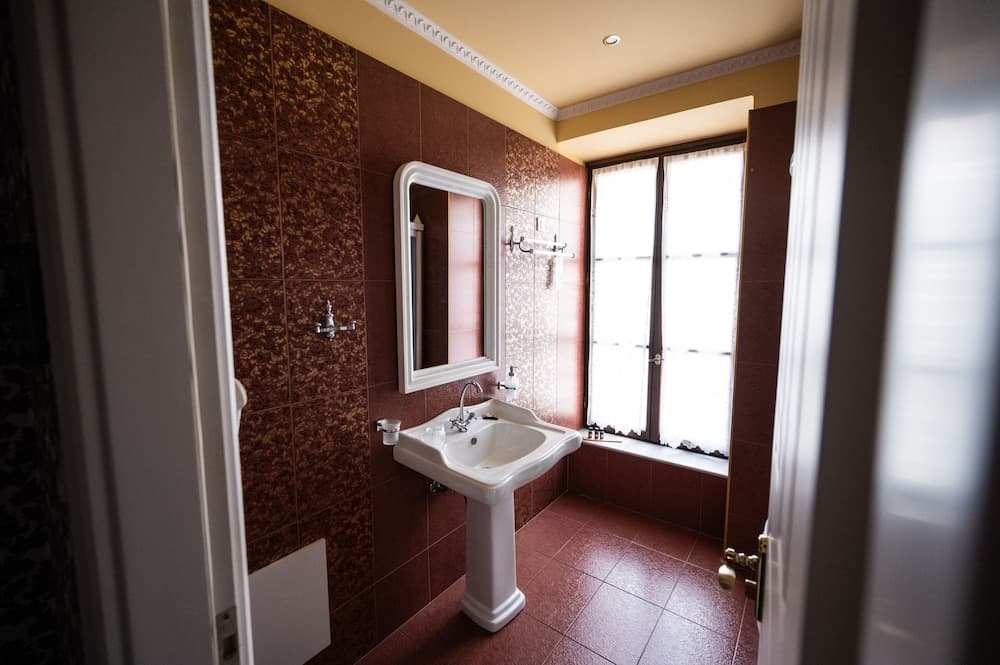 A cozy bathroom with warm-toned tiles, a white pedestal sink, and a bright window creates an inviting atmosphere, perfect for travelers seeking comfort in their accommodations.