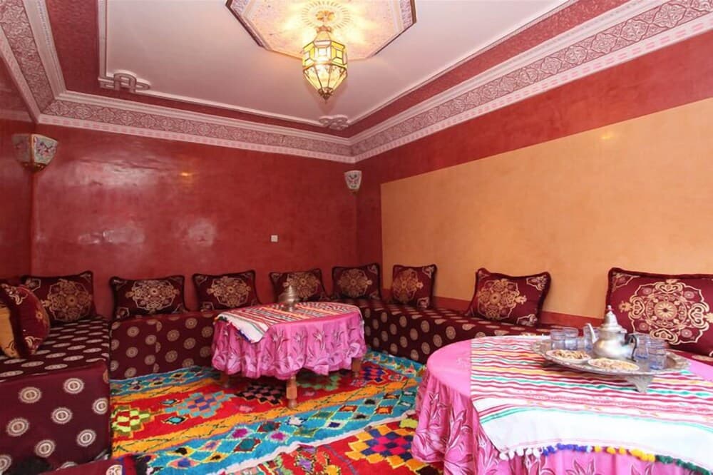 A cozy Moroccan-style lounge features vibrant red walls, ornate cushions, colorful rugs, and traditional tables set for tea, ideal for travelers seeking an authentic cultural experience.