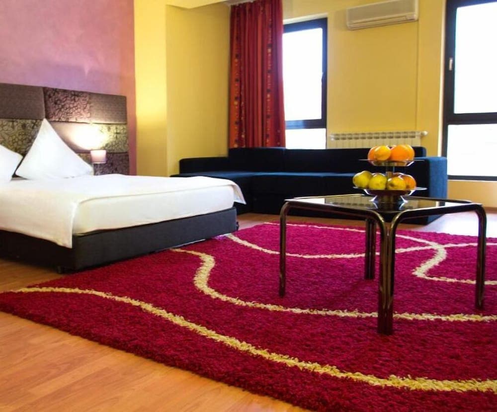 A cozy hotel room features a vibrant red rug, plush bed, modern sofa, and fruit-adorned coffee table, creating an inviting atmosphere ideal for travelers seeking comfort and style.