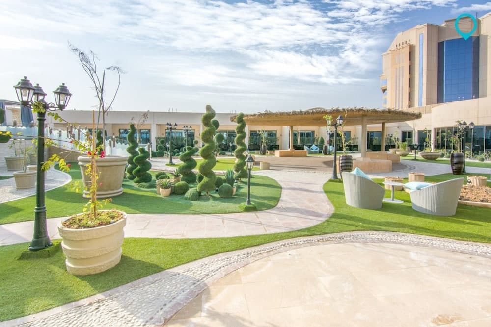 A serene outdoor courtyard features lush greenery, spiral topiaries, cozy seating areas, and elegant lampposts under a clear blue sky, ideal for leisurely strolls or relaxing getaways near modern buildings.