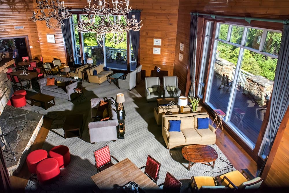 A cozy, rustic lodge lounge with wooden interiors, antler chandeliers, plush seating, and large windows overlooking lush greenery, ideal for travelers seeking relaxation in a serene mountain retreat.