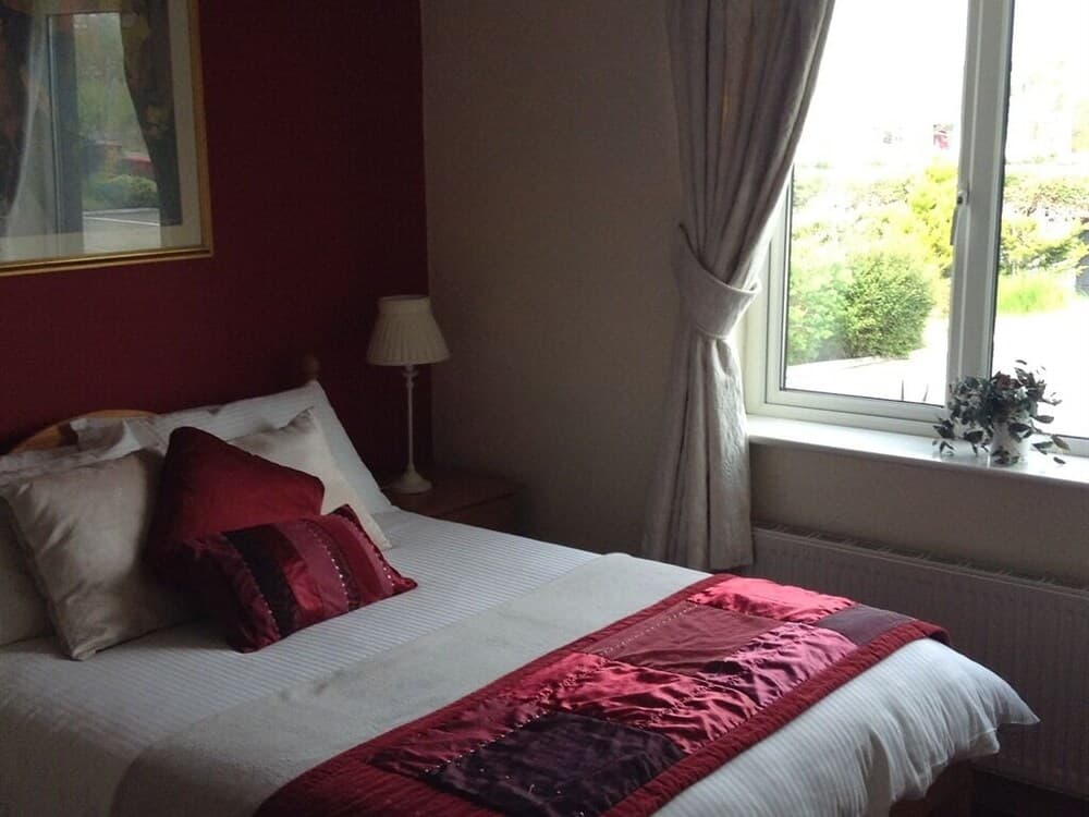 A cozy hotel room features a neatly made bed adorned with red and white bedding, a bedside lamp, and a window offering a serene view of greenery, ideal for travelers seeking comfort and relaxation.