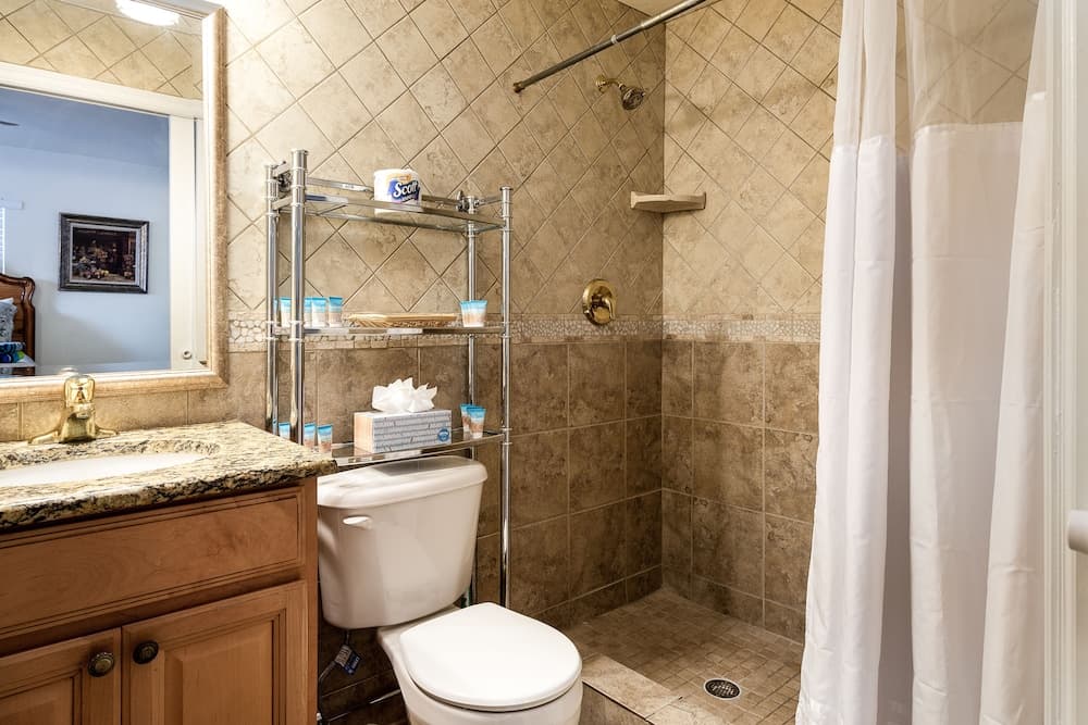 A cozy bathroom with beige tiled walls, a granite countertop vanity, a walk-in shower with a white curtain, and a chrome shelving unit stocked with toiletries, ideal for travelers seeking comfort and convenience.