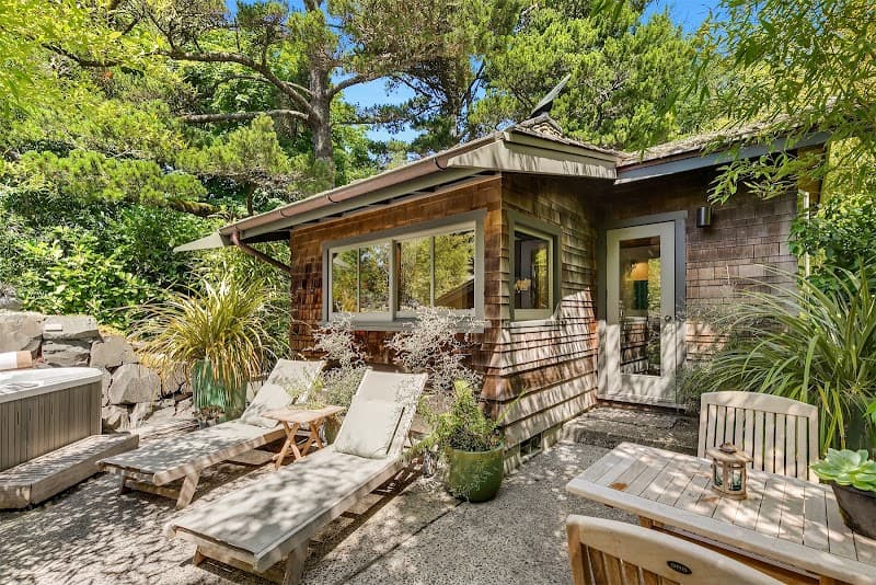 A serene coastal cabin surrounded by lush greenery features a cozy outdoor seating area with lounge chairs and a hot tub, perfect for a relaxing getaway in Manzanita, Oregon.