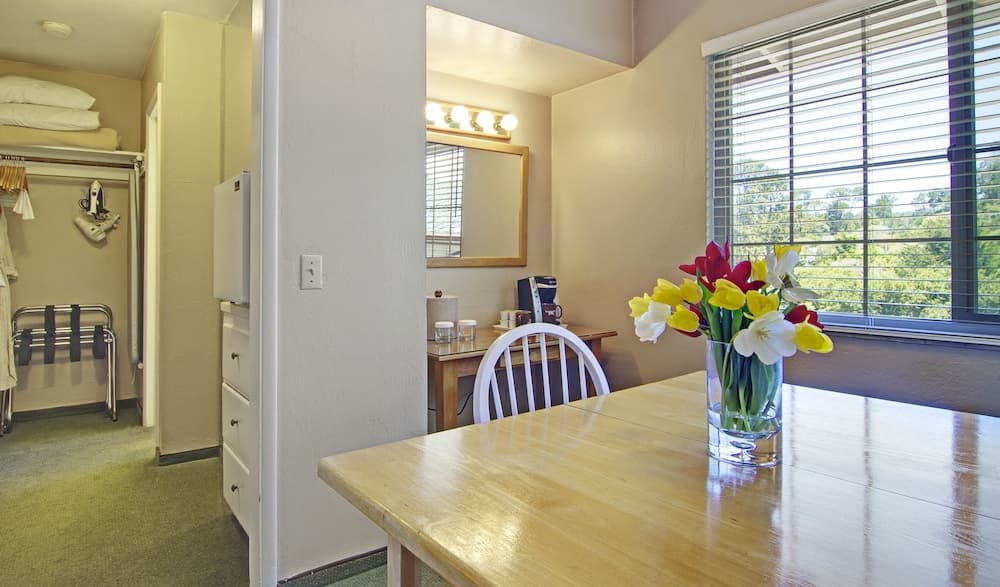 A cozy apartment dining area features a wooden table adorned with vibrant flowers, a bright window view of greenery, and a compact kitchenette, ideal for travelers seeking comfort and convenience in a homey setting.