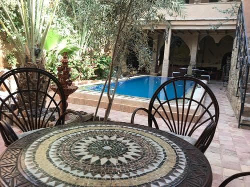 A serene courtyard with a mosaic table, wicker chairs, and a tranquil pool surrounded by lush greenery invites travelers to relax in a cozy, exotic retreat.