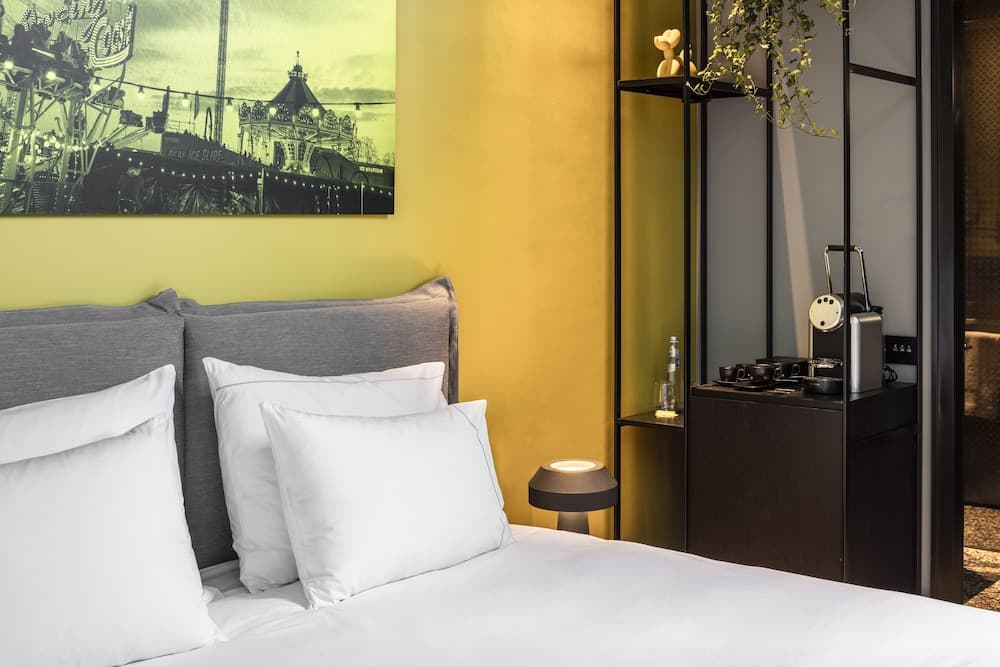 A cozy hotel room features a neatly made bed against a vibrant yellow wall adorned with a carnival-themed artwork, complemented by a modern shelving unit with a coffee station, ideal for travelers seeking comfort and convenience.