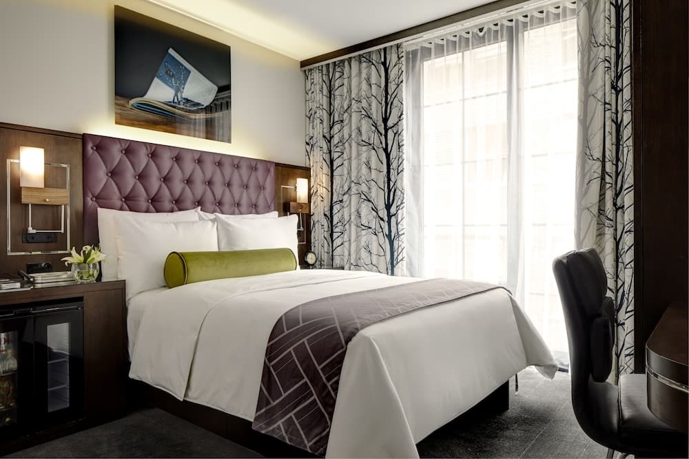 A cozy hotel room features a plush bed adorned with white linens and a green accent pillow, framed by tree-patterned curtains and modern bedside lamps, ideal for travelers seeking comfort and style.