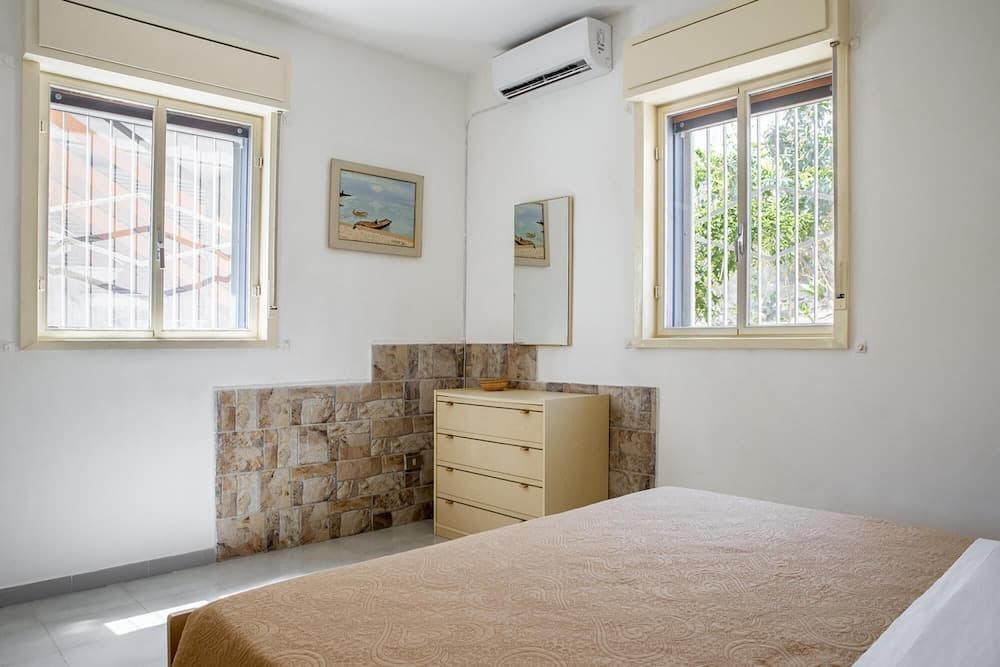 A cozy bedroom with a neatly made bed, stone-tiled accent wall, two windows offering views of greenery and buildings, and a dresser adorned with framed artwork, ideal for travelers seeking a serene and comfortable stay.