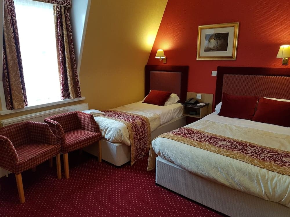 A cozy hotel room with vibrant red walls, two neatly made beds adorned with patterned throws, a window with elegant curtains, and matching armchairs, ideal for travelers seeking comfort and relaxation.