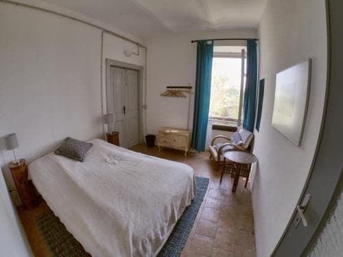 A cozy bedroom with terracotta flooring, white walls, and a large window draped in teal curtains offers a serene retreat, featuring a neatly made bed, vintage furniture, and natural light, ideal for travelers seeking a peaceful accommodation experience.