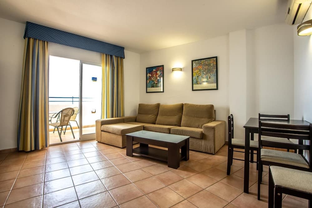 A cozy apartment living room with tiled floors, a beige sofa, dining area, colorful curtains, and a balcony view, ideal for travelers seeking comfortable accommodations near the sea.