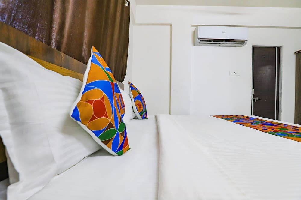 A cozy hotel room features a neatly made bed adorned with colorful pillows and a vibrant runner, set against a minimalist backdrop with an air conditioner and wooden accents, ideal for travelers seeking comfort and simplicity.