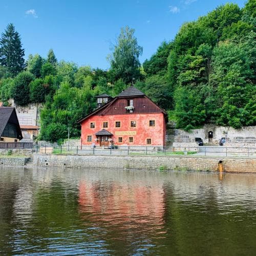 A charming red building nestled by a serene riverbank under lush greenery offers a picturesque setting for tranquil getaways, ideal for travelers seeking scenic relaxation spots.