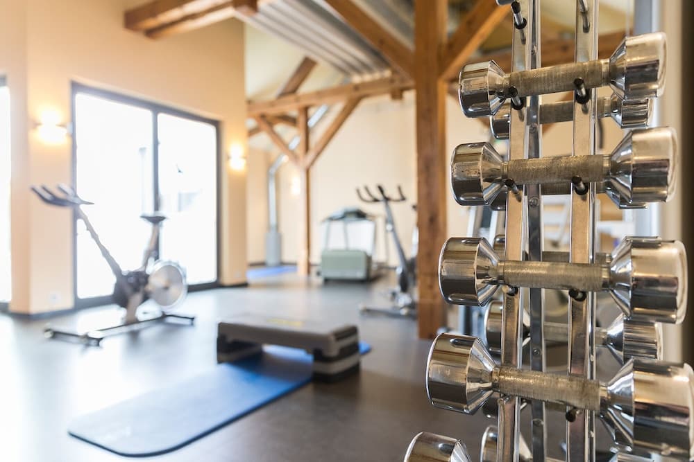 A well-lit gym with wooden beams features a rack of dumbbells in the foreground, stationary bikes, and exercise mats, creating an inviting atmosphere for fitness enthusiasts seeking a serene workout space.