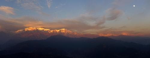 A breathtaking sunset illuminates snow-capped mountains under a serene sky with scattered clouds and a visible moon, ideal for adventure seekers exploring high-altitude landscapes.