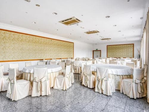 A spacious banquet hall with elegant white tablecloths and chairs set up for events, featuring patterned walls and a polished floor, ideal for wedding receptions or corporate gatherings.