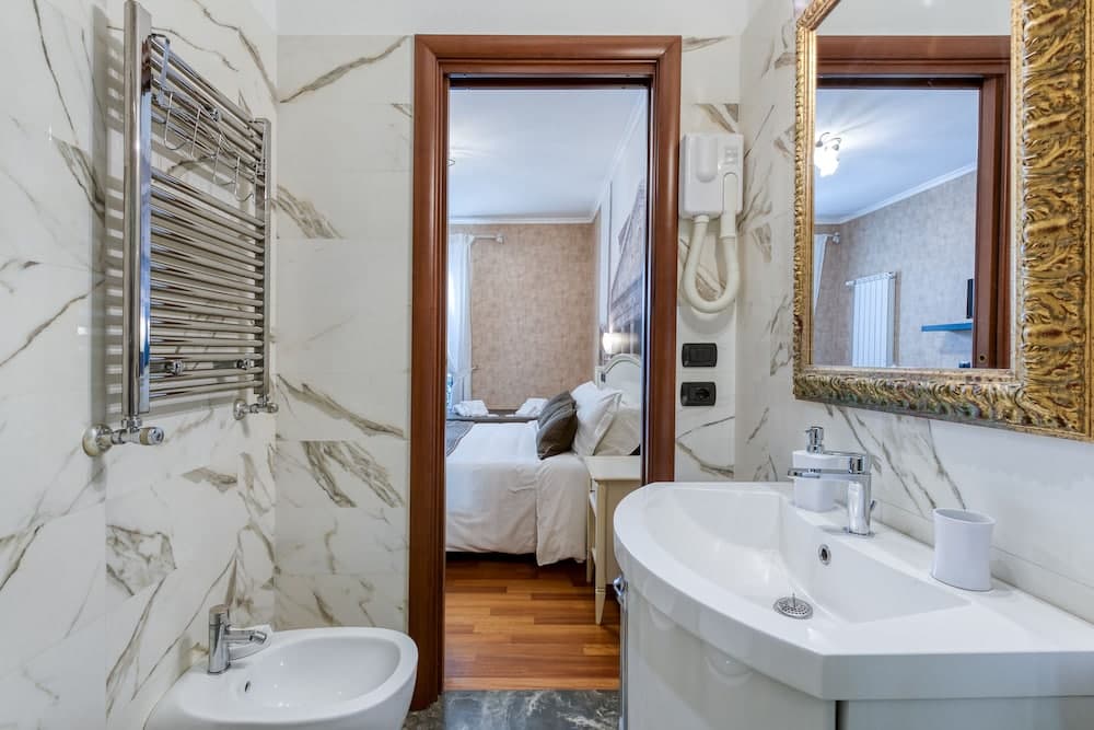 A luxurious bathroom with marble walls, a bidet, and an ornate mirror seamlessly connects to a cozy bedroom through a wooden doorway, ideal for travelers seeking comfort and elegance in their accommodations.