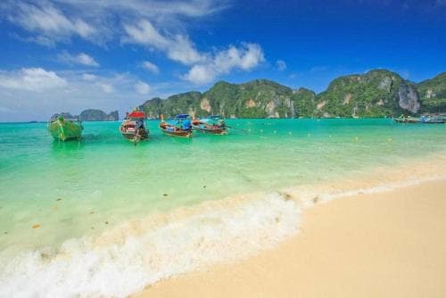 A serene tropical beach with turquoise waters, colorful longtail boats, and lush green mountains in the background offers an idyllic escape for travelers seeking relaxation and adventure.