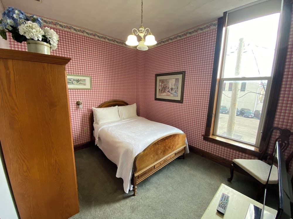 A cozy hotel room with red checkered wallpaper, a wooden bed, a chandelier, and a window view of parked cars, ideal for travelers seeking a quaint and comfortable stay.
