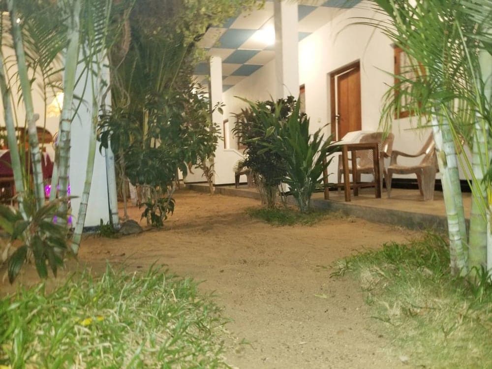 A tranquil evening scene at a tropical resort features a sandy path lined with lush greenery and bamboo plants leading to a cozy seating area outside a well-lit building, ideal for travelers seeking relaxation and nature immersion.