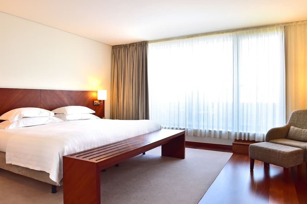 A serene hotel room features a cozy bed with crisp white linens, a wooden bench, soft lighting, and large windows draped with sheer curtains, ideal for travelers seeking comfort and relaxation.
