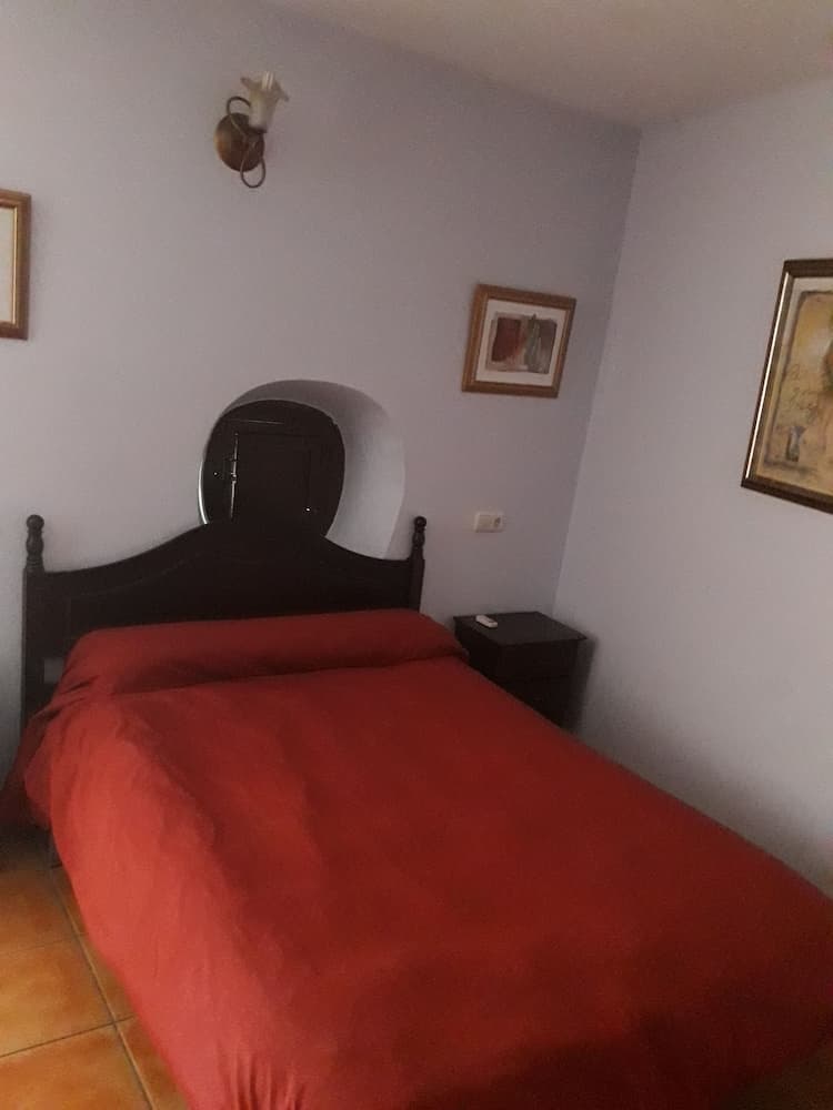 A cozy bedroom with a red bedspread, wooden furniture, framed artwork, and a wall-mounted lamp offers a serene retreat for travelers seeking a comfortable stay.