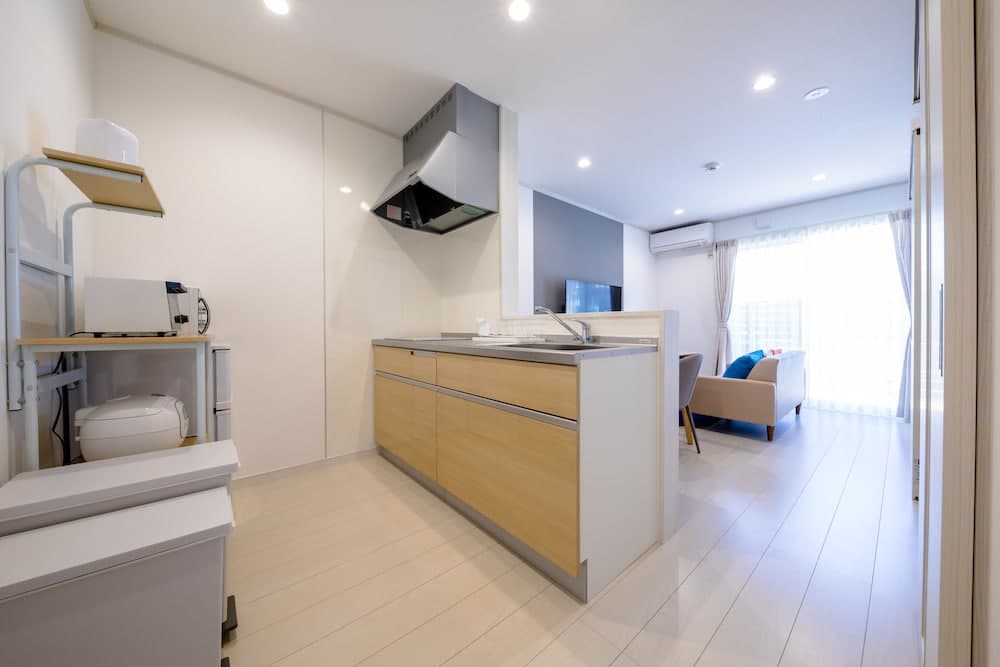 A modern, minimalist apartment kitchen seamlessly blends into an open living area, featuring wooden cabinets, a sleek countertop, and ample natural light from large windows, ideal for travelers seeking cozy and functional accommodations.